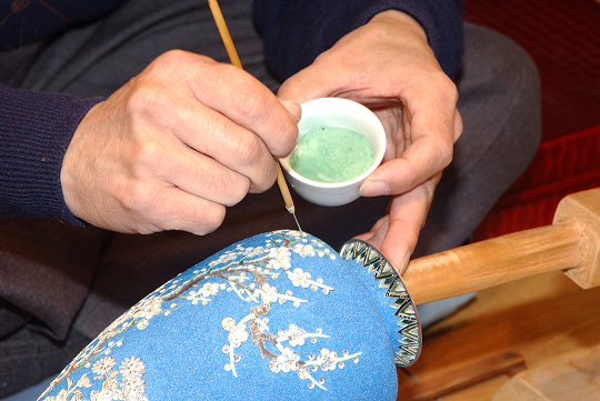 木綿針を軸の先端に取り付けた「針ボセ」や、極細の面相筆（めんそうふで）で釉薬を重ねる。緻密な指先の制御と経験値が発色を左右する、尾張七宝の心が宿る瞬間。